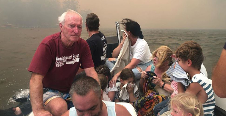 Boat filled with people on lake with fire raging behind them