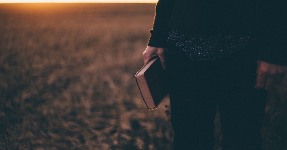person standing in a field at sunset holding a bible