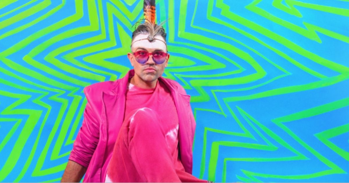 mitch tambo wearing a bright pink tracksuit and pink sunglasses on a crazy green and blue background