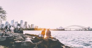 Two people sitting and standing at shore watching the sunset near the Sydney Opera House in panorama photography