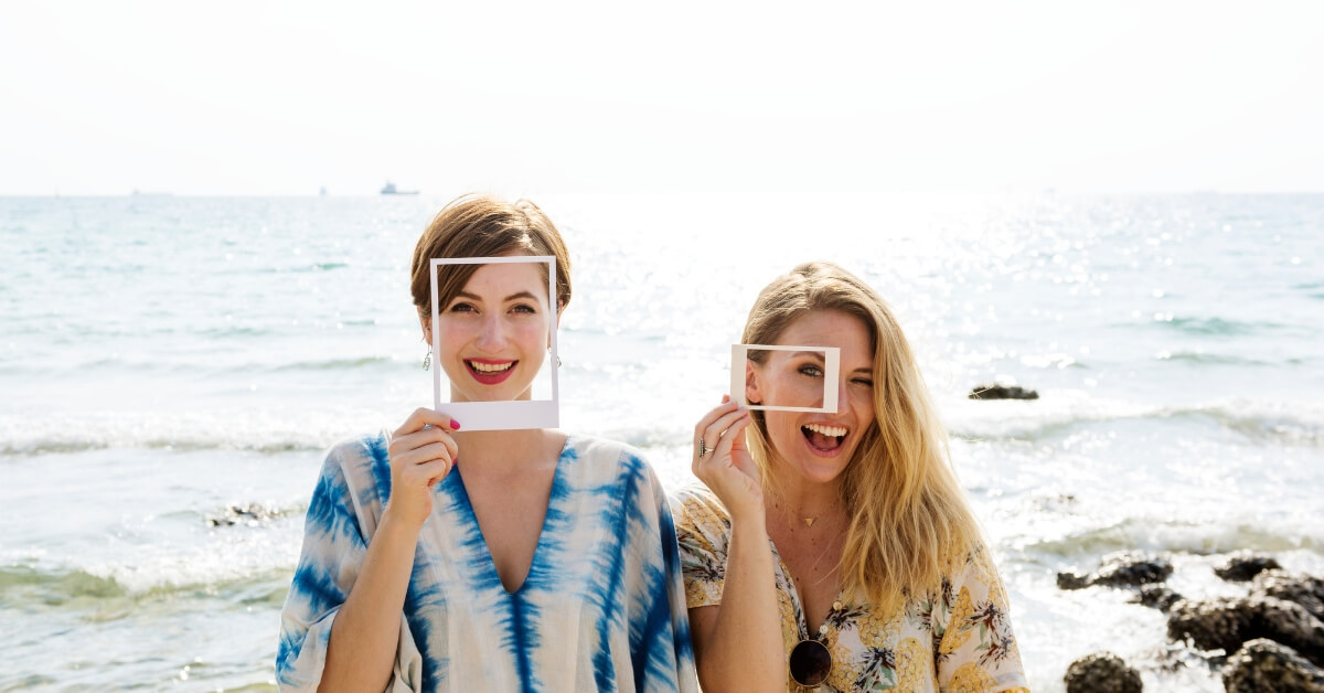 two women standing in from of ocean holding up Polaroid frames to their faces