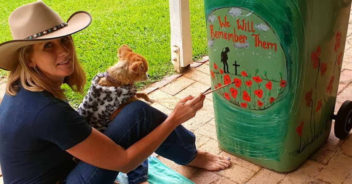Woman in cowboy hat with dog on lap paints Anzac Memorial picture on her wheelie bin