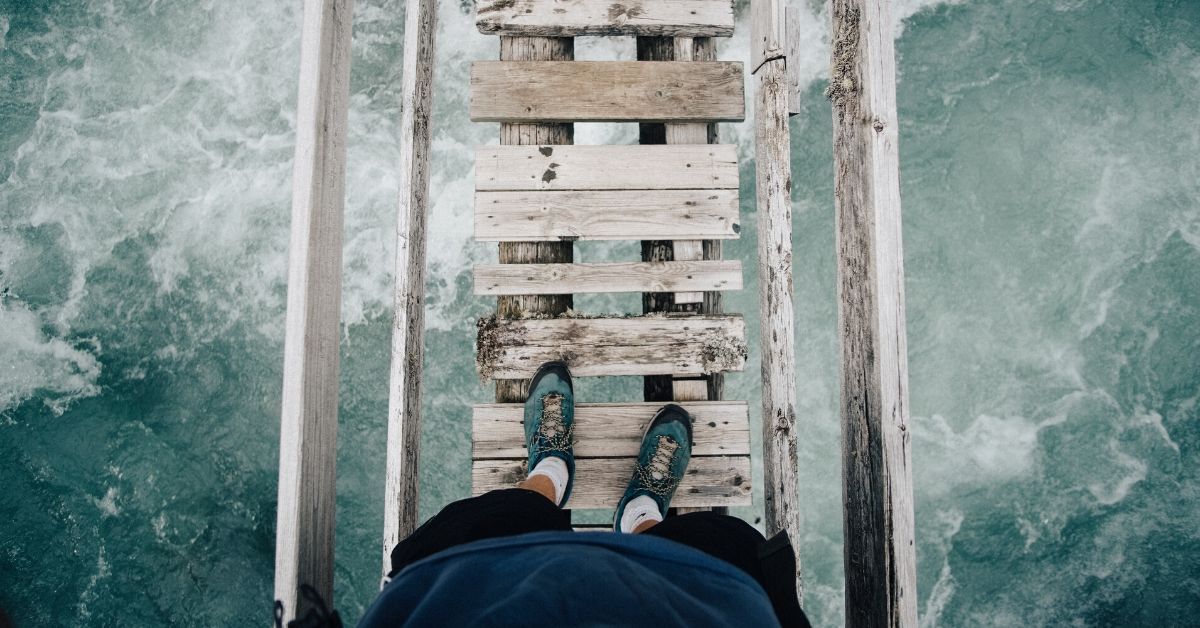 photo of a person's legs standing on a rickety bridge with rapid water underneath