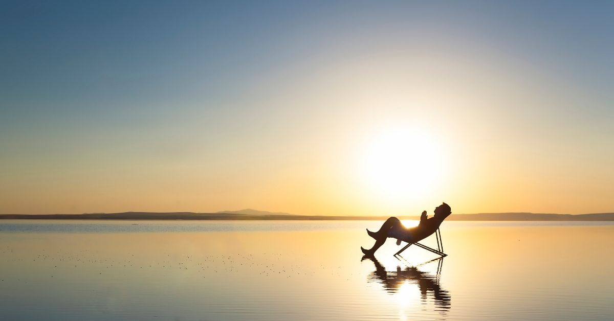 photo shows a silhouette of a lone man reclining in a deck chair on a beach at sunrise