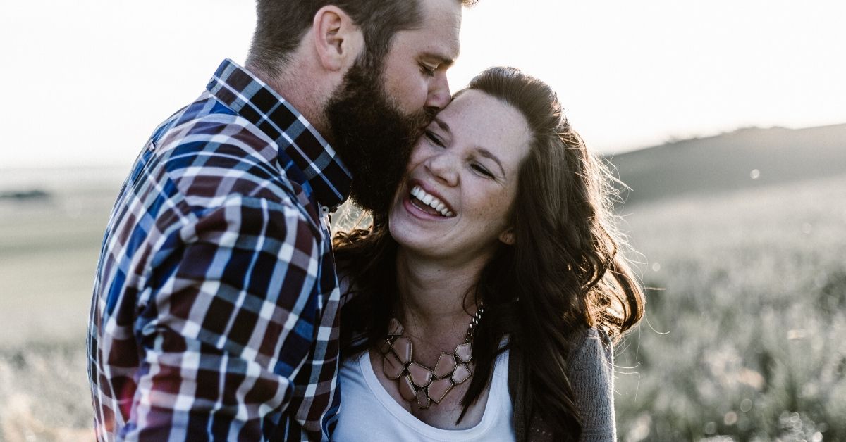 photo depicts a man kissing a woman on the side of her head as she laughs happily
