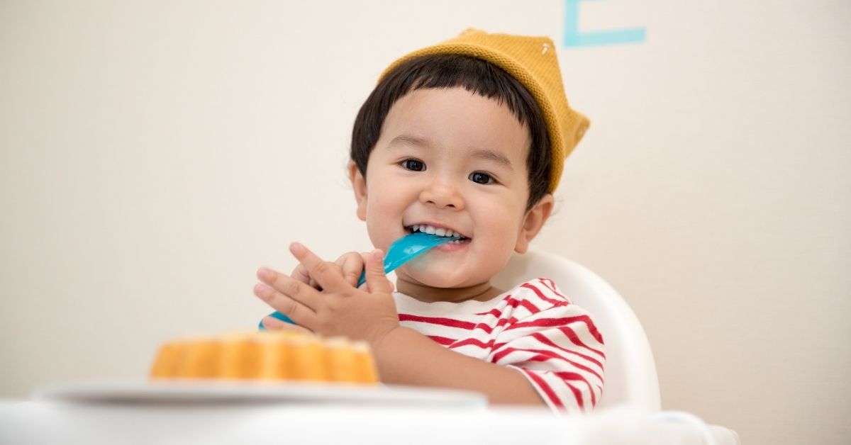 a toddler smiling at the camera with a spoon in his mouth and cake in front of him