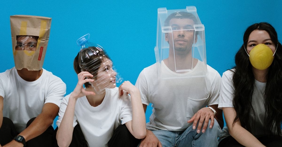 a conceptual photo of people wearing white shirts wearing masks made out of products such as a paper bag, bucket and cut in half plastic bottle posing in front of a blue backdrop