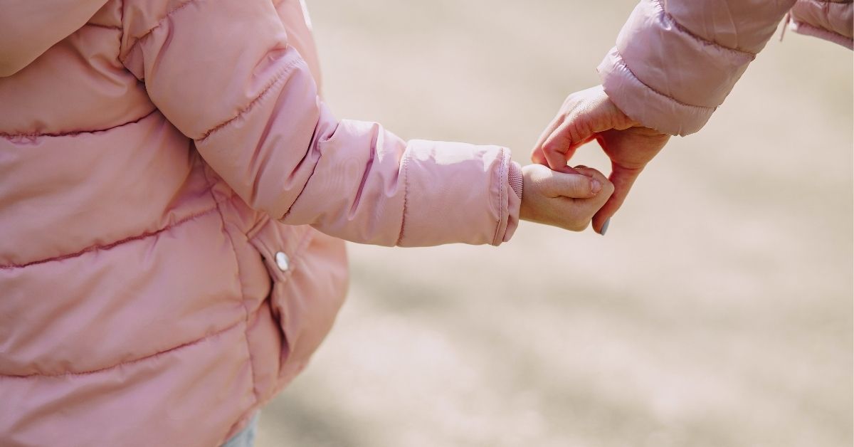a young girl wearing a pink jacket holds her mother's hand as they walk