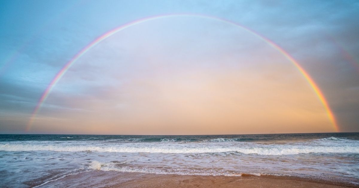 a large rainbow stretchs across the sky over a beach at sunset